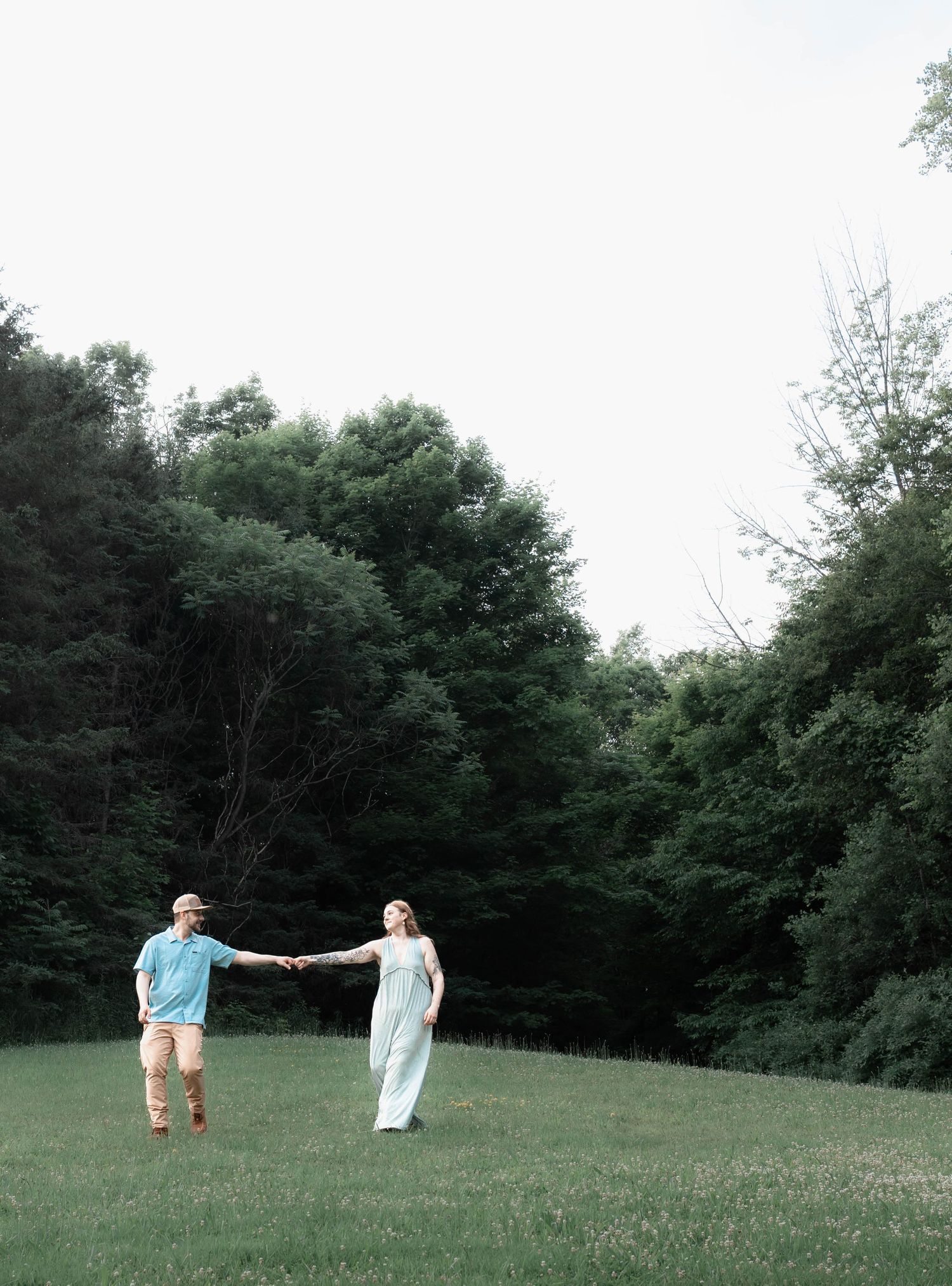 Two people standing together in a grassy field surrounded by green trees on a sunny summer day.