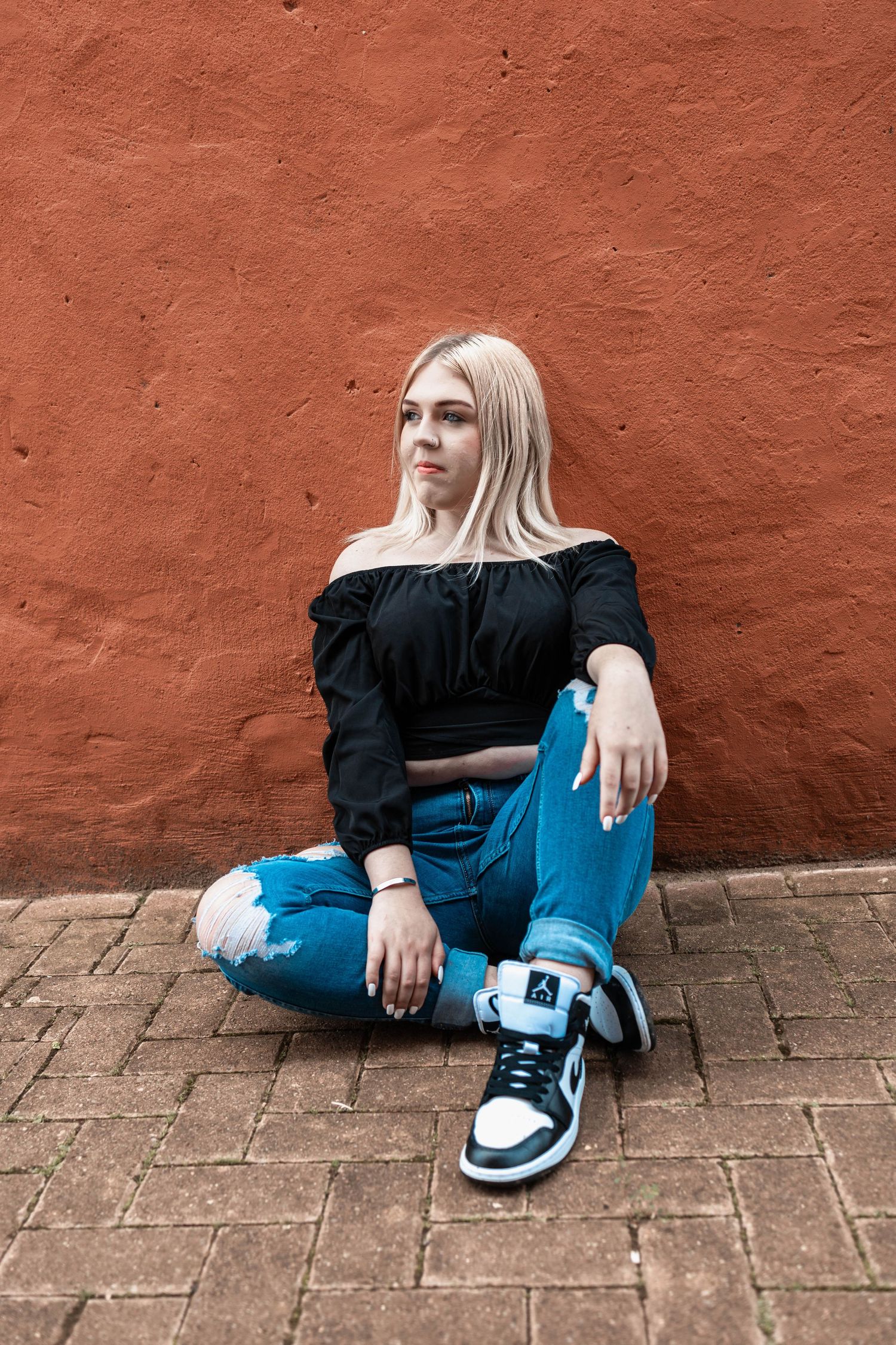Person in black top and ripped jeans sitting against terracotta wall wearing sneakers.
