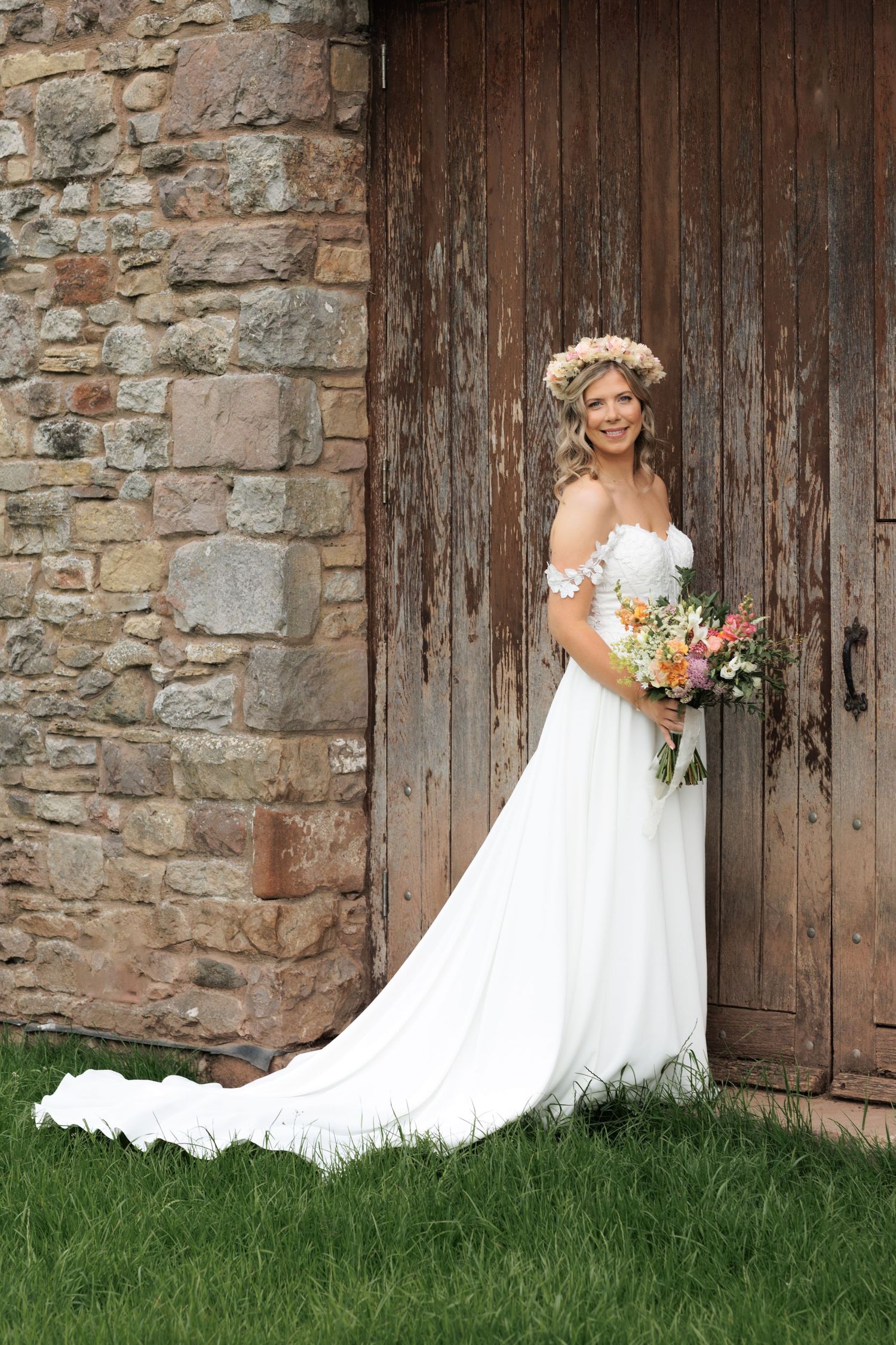 A bride in white poses with flowers against a rustic wooden door and stone wall.