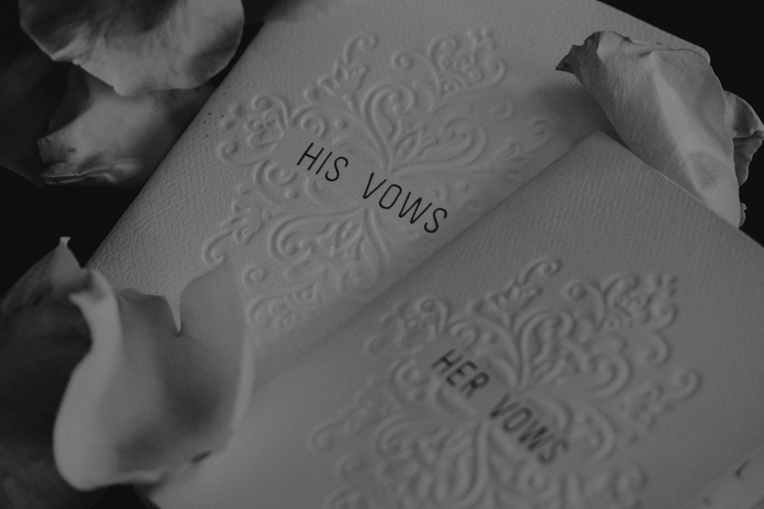 A wedding vow book with embossed text reading 'His Vows' and 'Her Vows' with white rose petals surrounding it.
