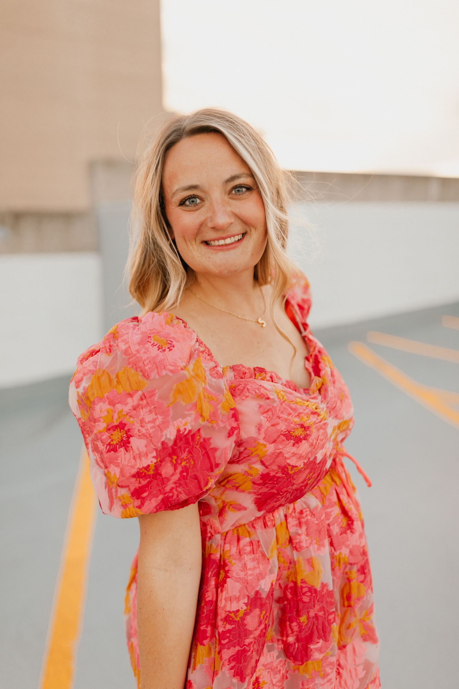 Woman in vibrant pink floral dress with puff sleeves smiles in a parking structure at sunset on a rooftop in Lexington, Kentucky.