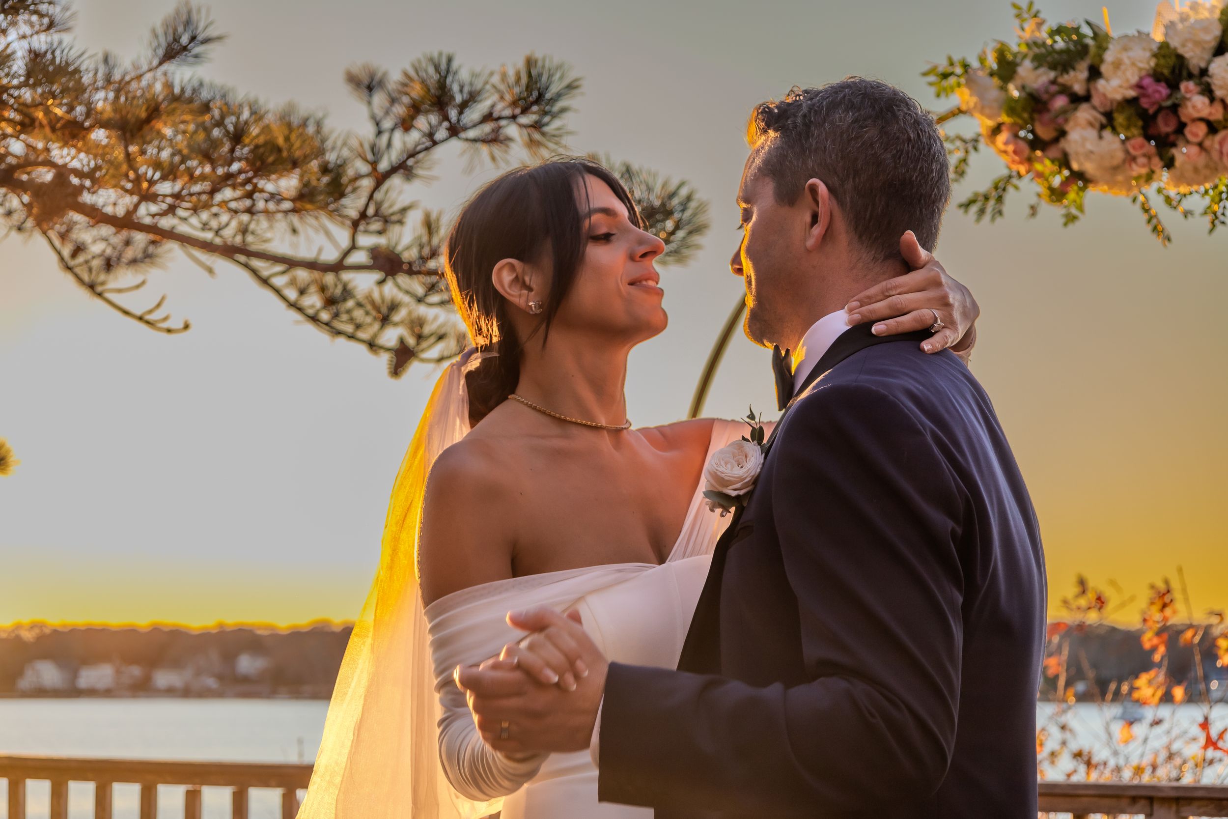 A bride and groom share an intimate moment at sunset on a waterfront balcony with golden light illuminating the scene.