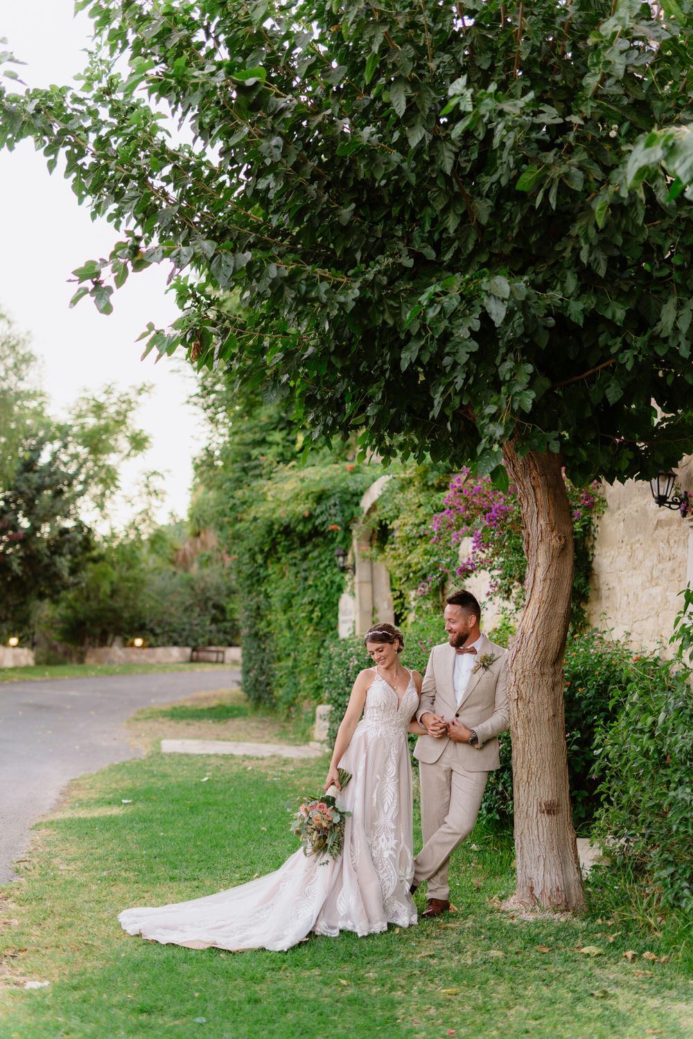 A couple poses beneath leafy trees in formal wedding attire on a garden path.