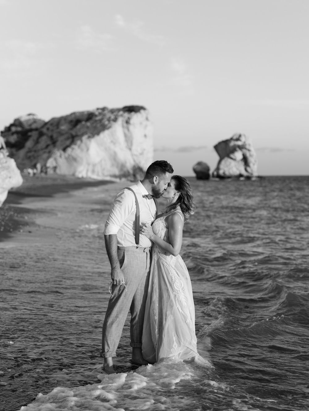A black and white photo of a couple embracing at the beach with dramatic cliffs in the background. rock of aphrodie