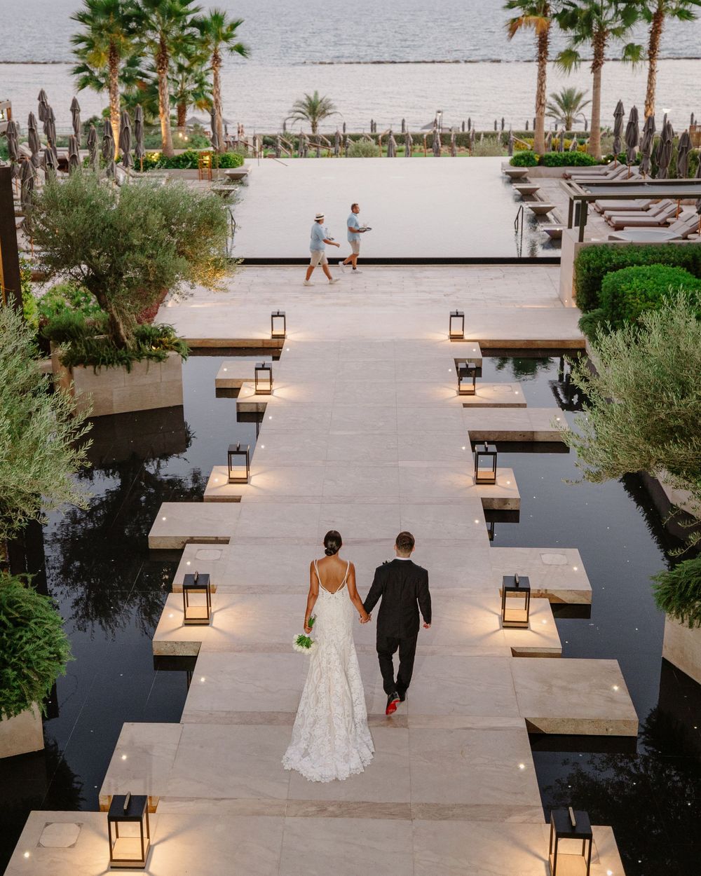 Couple walks down illuminated pathway lined with lanterns and palm trees at sunset.