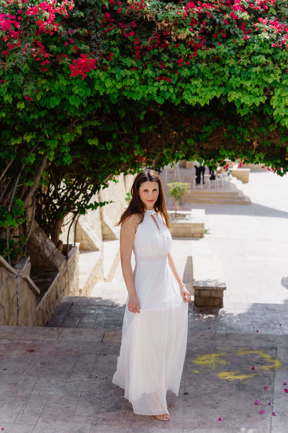 Bride in flowing white halter dress poses beneath archway of vibrant red flowers.