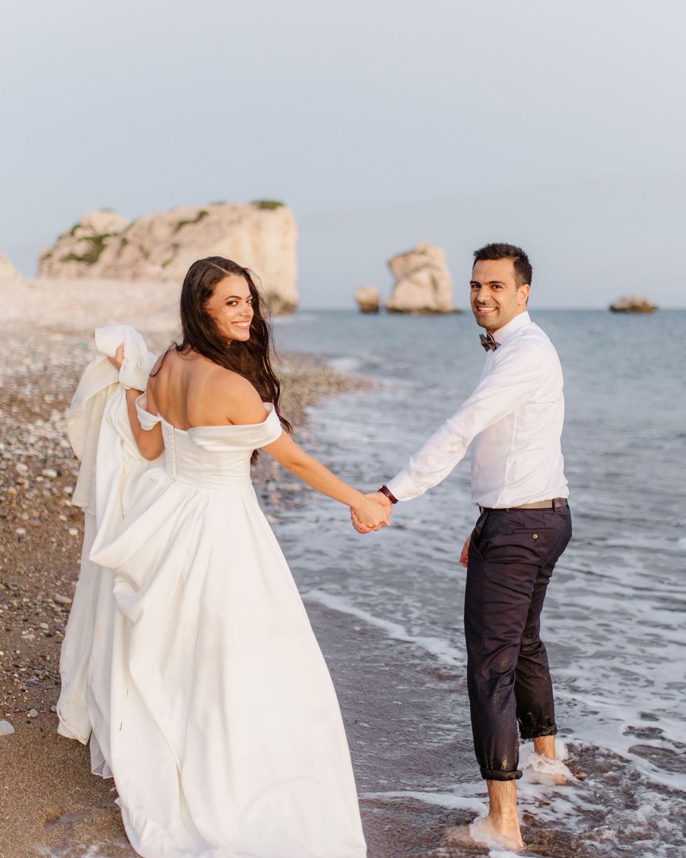 Bride and groom walk barefoot along the shore at the Rock of Aphrodite, laughing and holding hands in a joyful seaside moment.