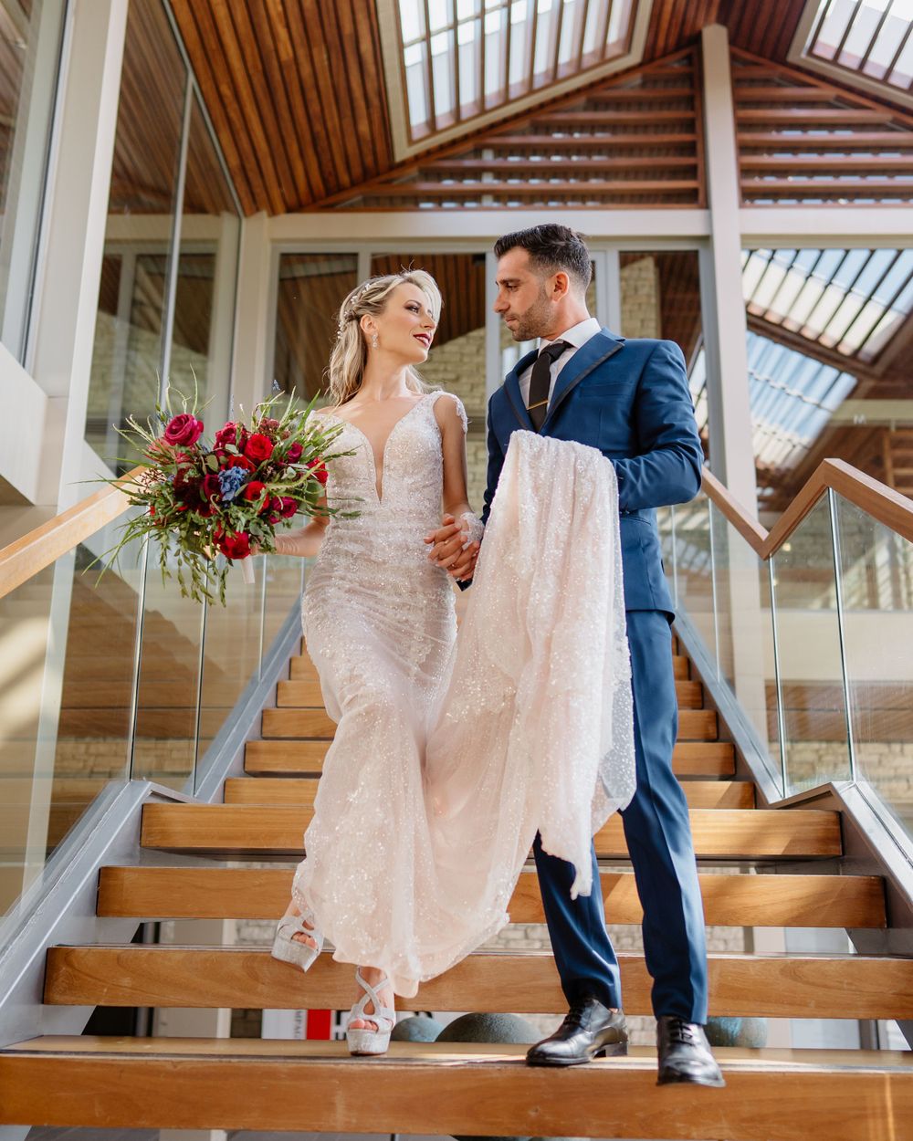 A bride and stands on wooden stairs in a bright venue, featuring a navy suit and white wedding gown with red bouquet. minthis hills