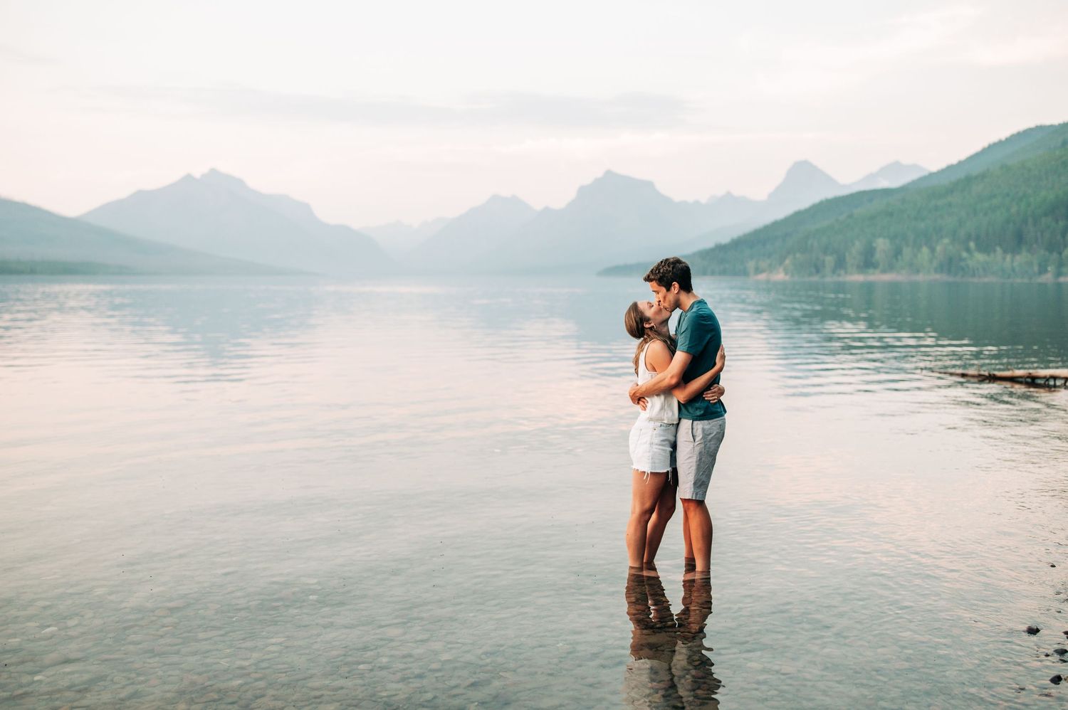 A couple embraces while standing in the shallow waters of a lake surrounded by mountains at sunset.