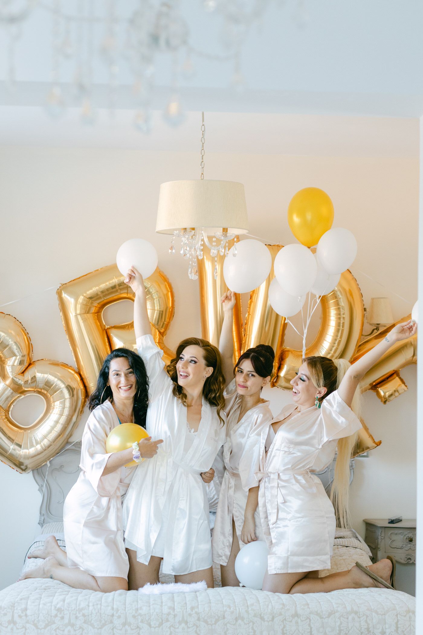 Friends in white robes celebrate on a bed with gold balloon numbers and festive decorations for a special occasion.