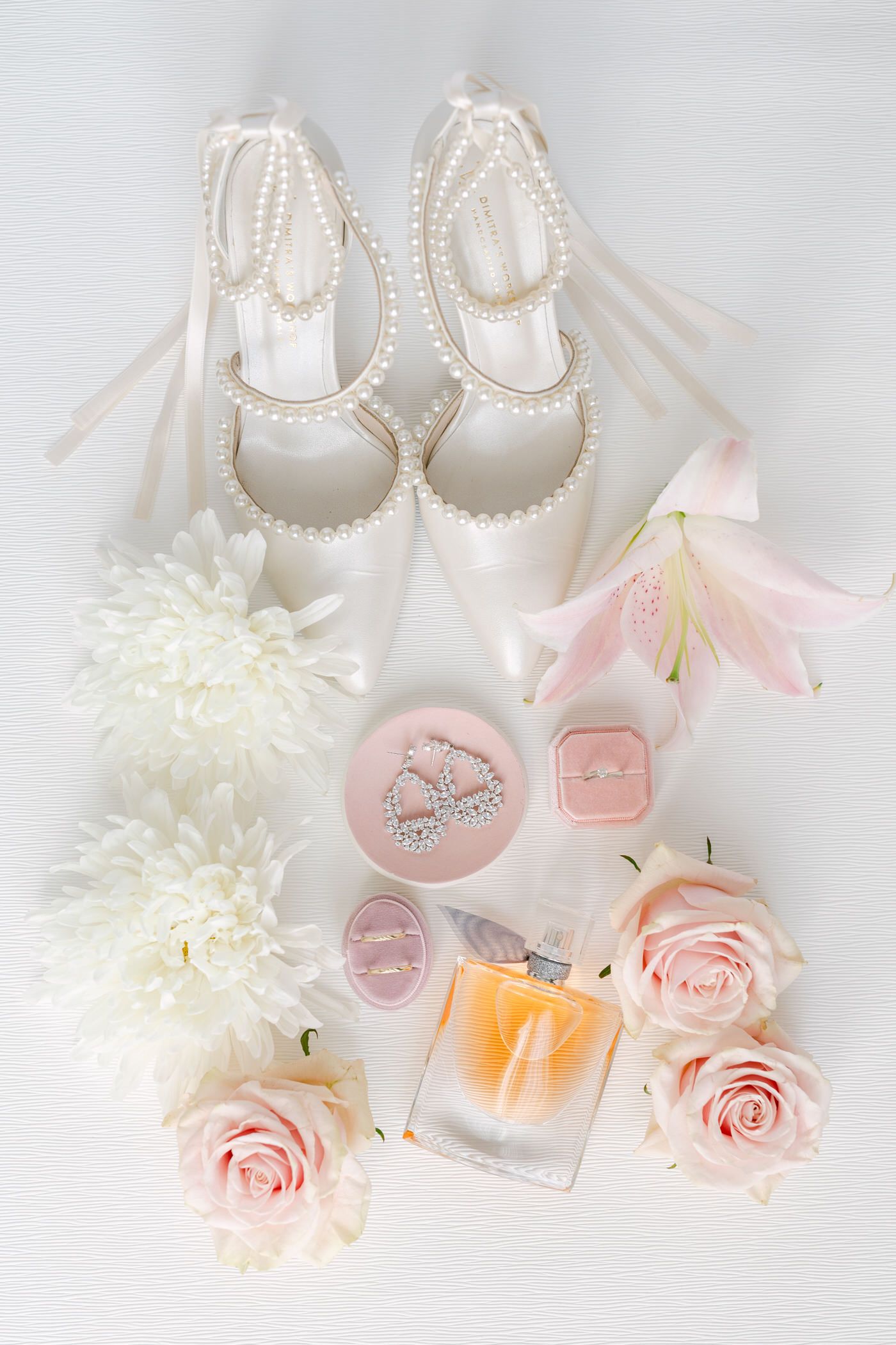 Elegant white bridal shoes with pearl details displayed with pink roses, perfume bottle and delicate pastel wedding accessories.