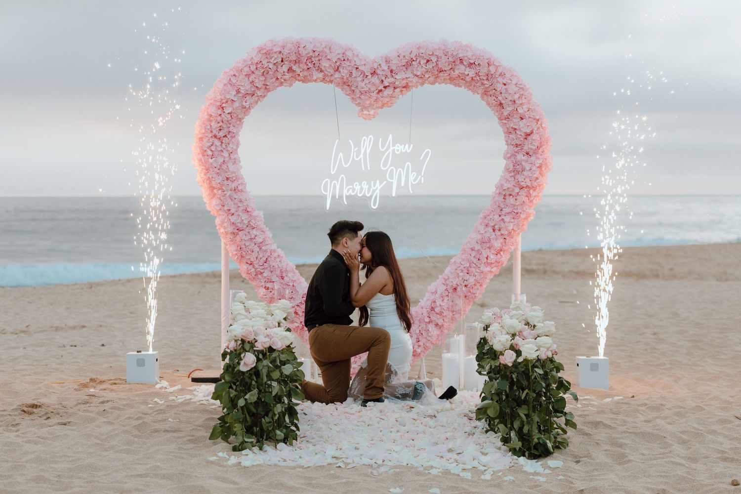 A romantic beach proposal setup with a pink heart-shaped arch, cold spark fireworks and floral arrangements on the sand.