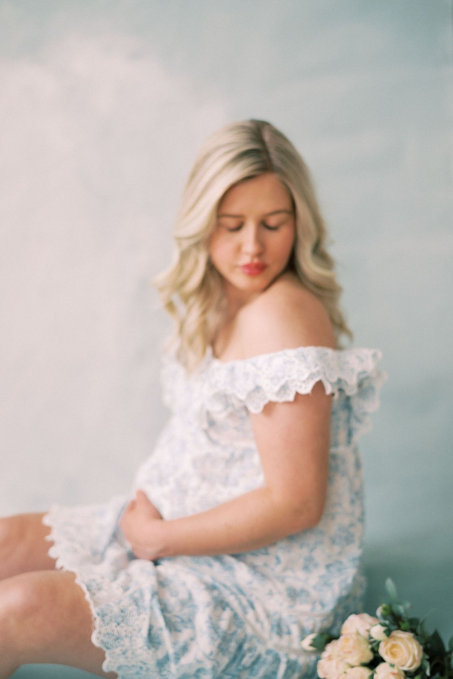 A dreamy portrait of someone in a delicate white lace dress sitting with cream-colored roses against a soft blue backdrop.