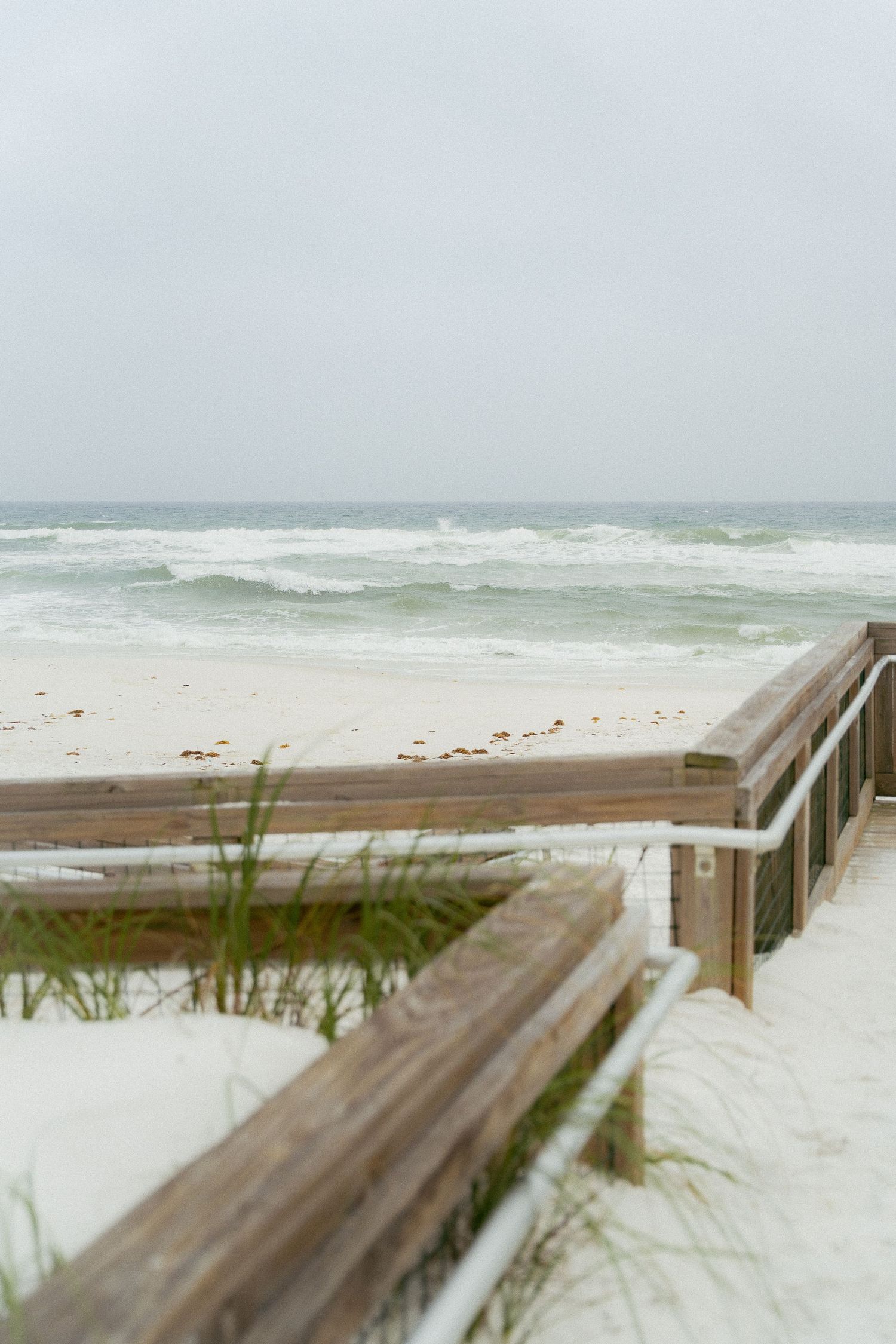Wooden boardwalk leads down to a stormy beach with white-capped waves and misty gray skies over the ocean.