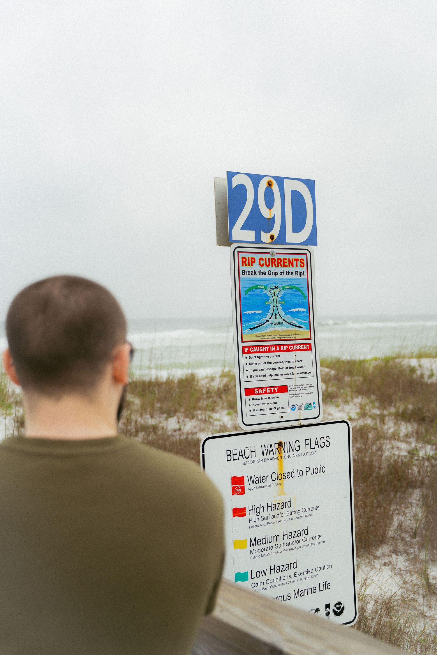 Person views beach warning sign displaying surf conditions and flag system on cloudy coastal day.