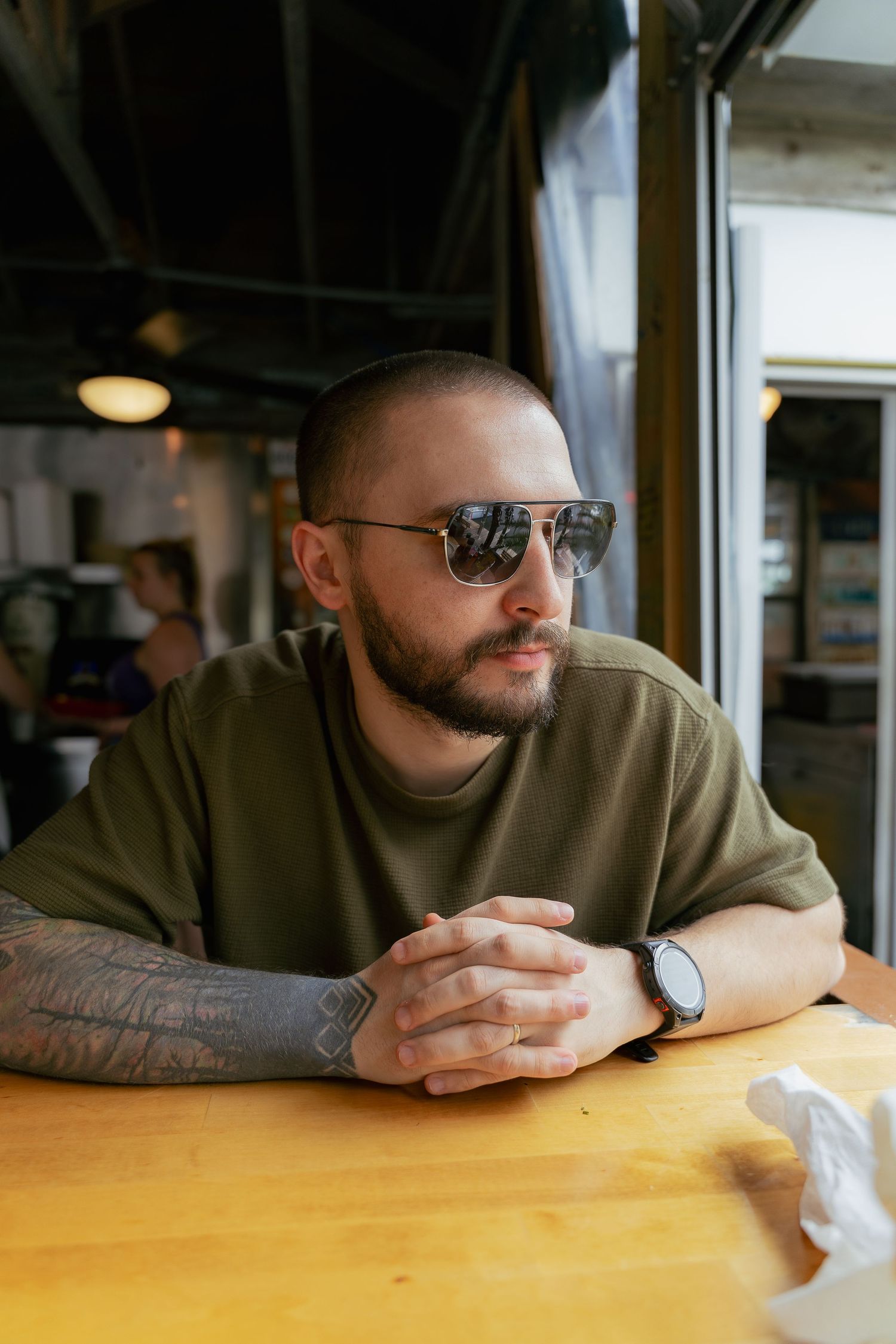 Person in sunglasses with a beard wearing olive sweater sits at wooden table in coffee shop, showing tattooed arm.