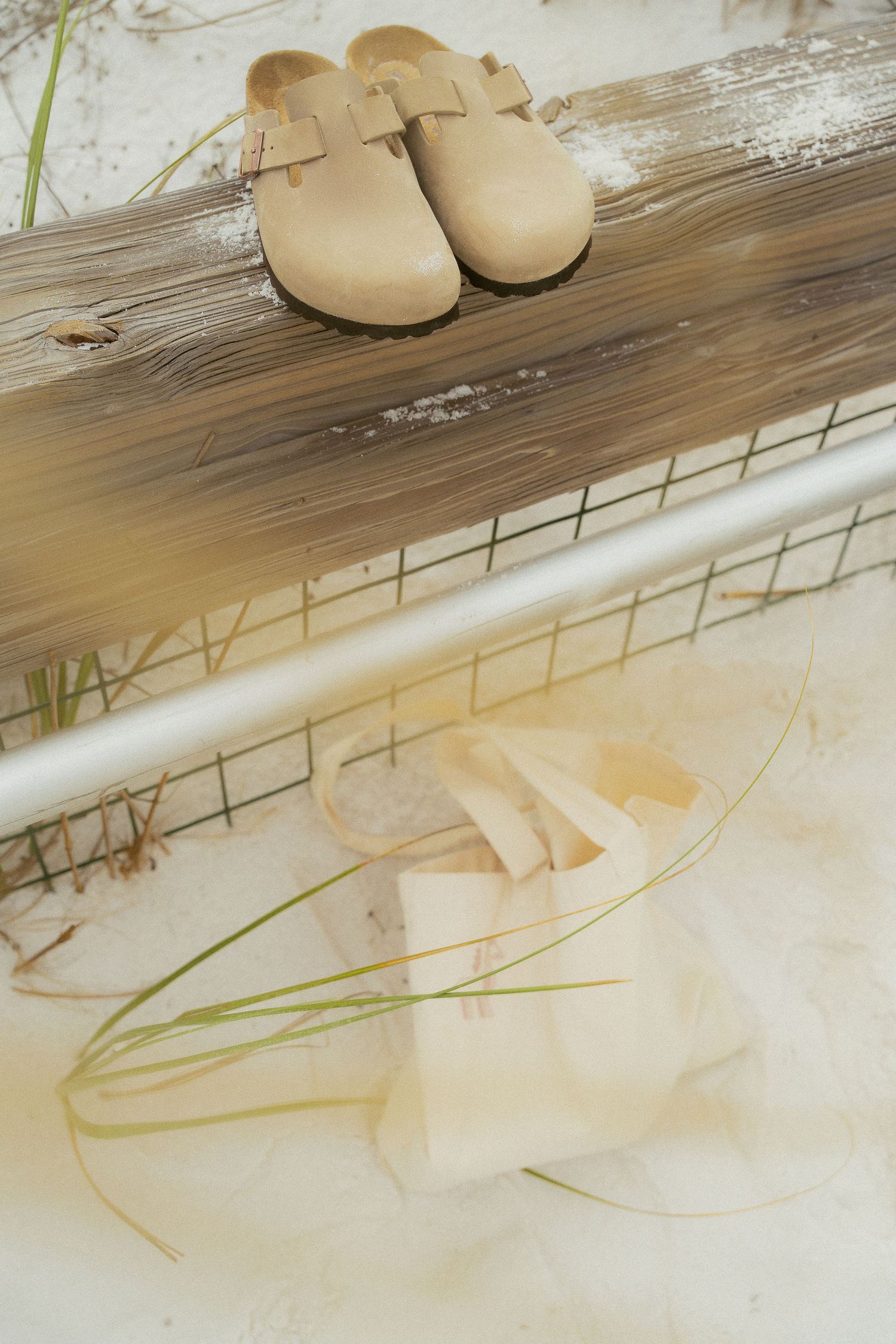 A pair of beige leather clogs sits on a rustic wooden surface with soft natural lighting and grass elements.