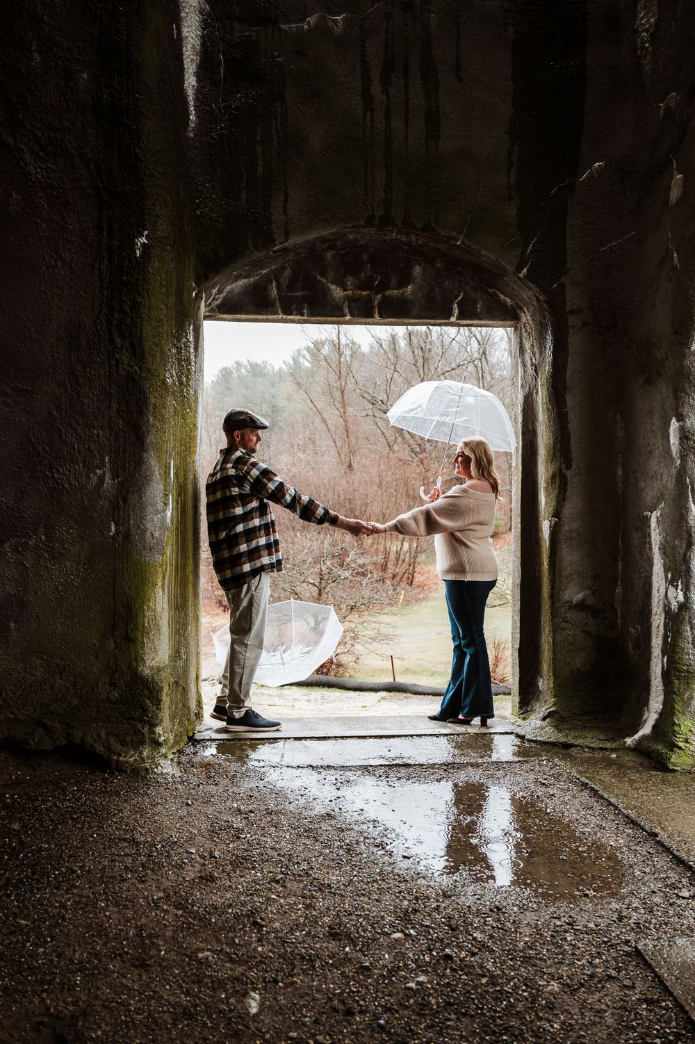 Two figures stand in silhouette within a stone archway tunnel while one holds a white umbrella on a rainy day.