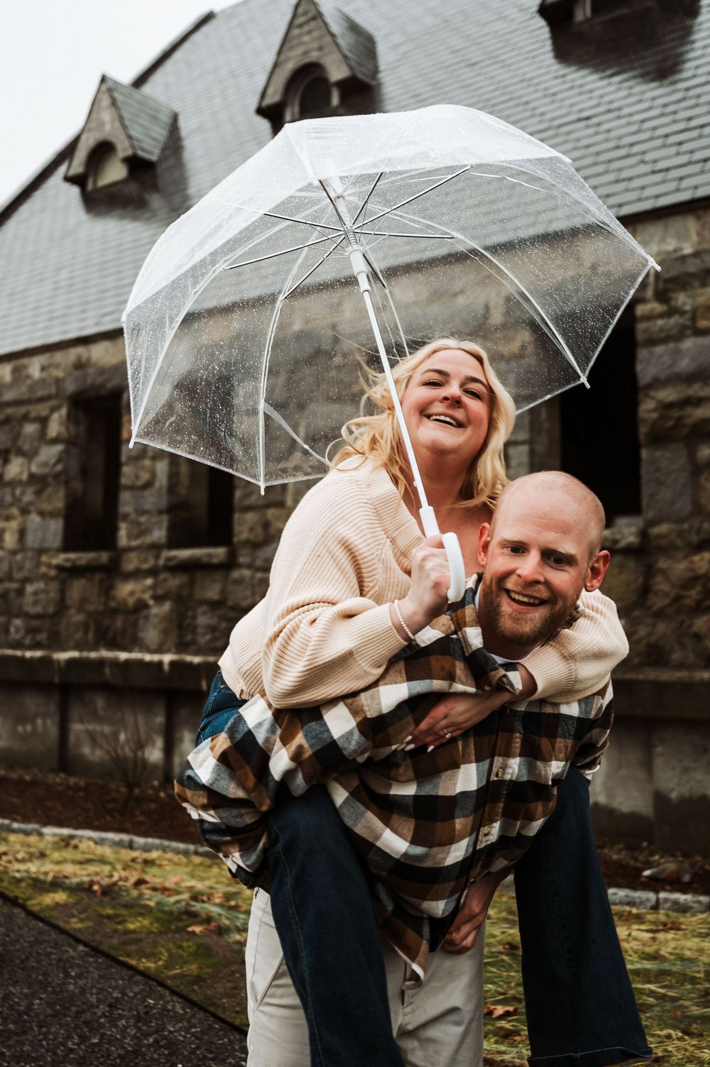 A couple shares a joyful moment under a clear umbrella outside a stone church on a rainy day.