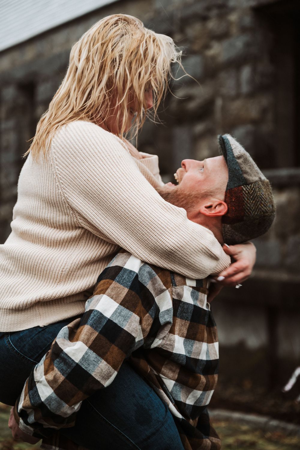 Couple in casual sweater and plaid shirt sharing an intimate emotional moment outdoors against stone wall background.