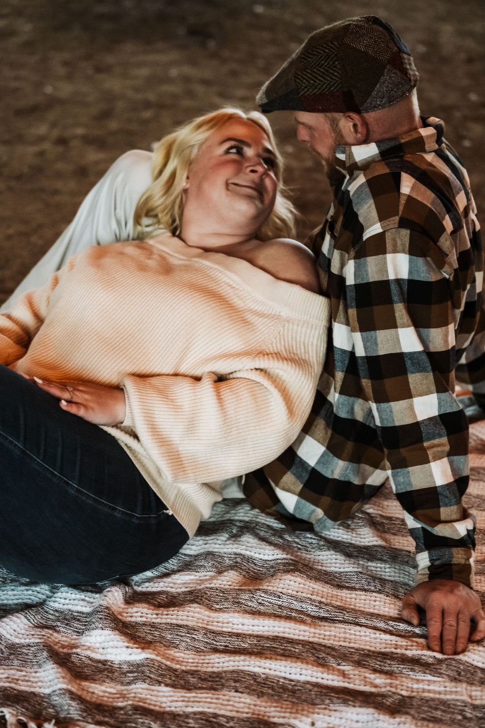 A couple shares a romantic moment while relaxing on a cozy blanket, wearing casual sweaters and plaid.