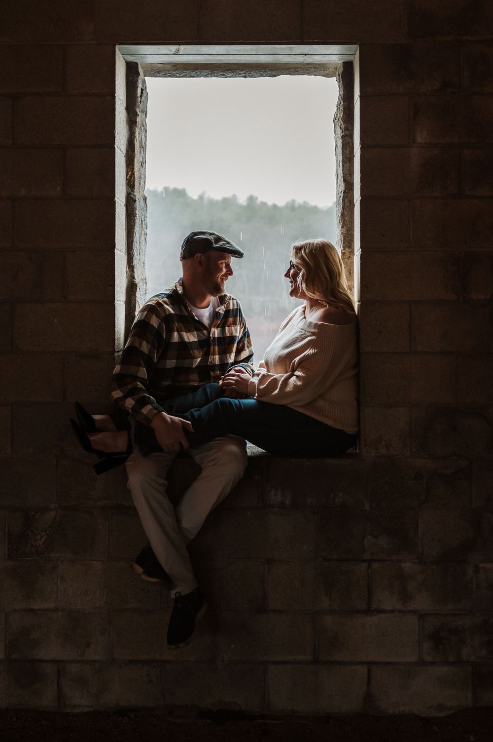 A couple sits together in a window frame silhouetted against misty mountains during golden hour.
