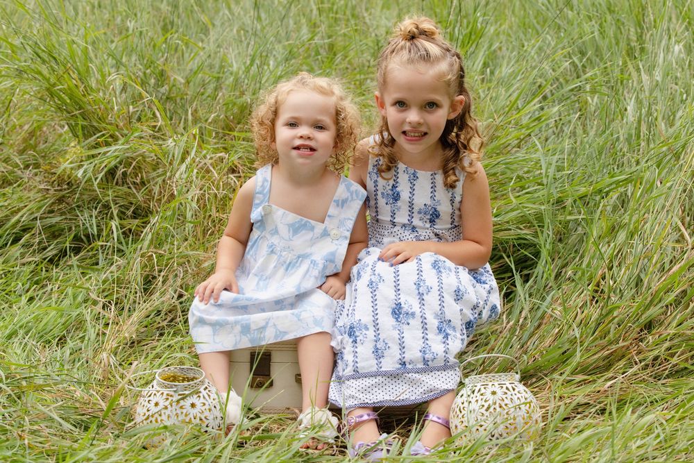 Children in floral dresses play together in a sunny meadow of tall grass swaying in the breeze.