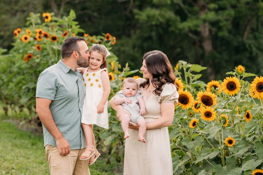 Family stands together in a garden with tall blooming sunflowers on a summer afternoon.