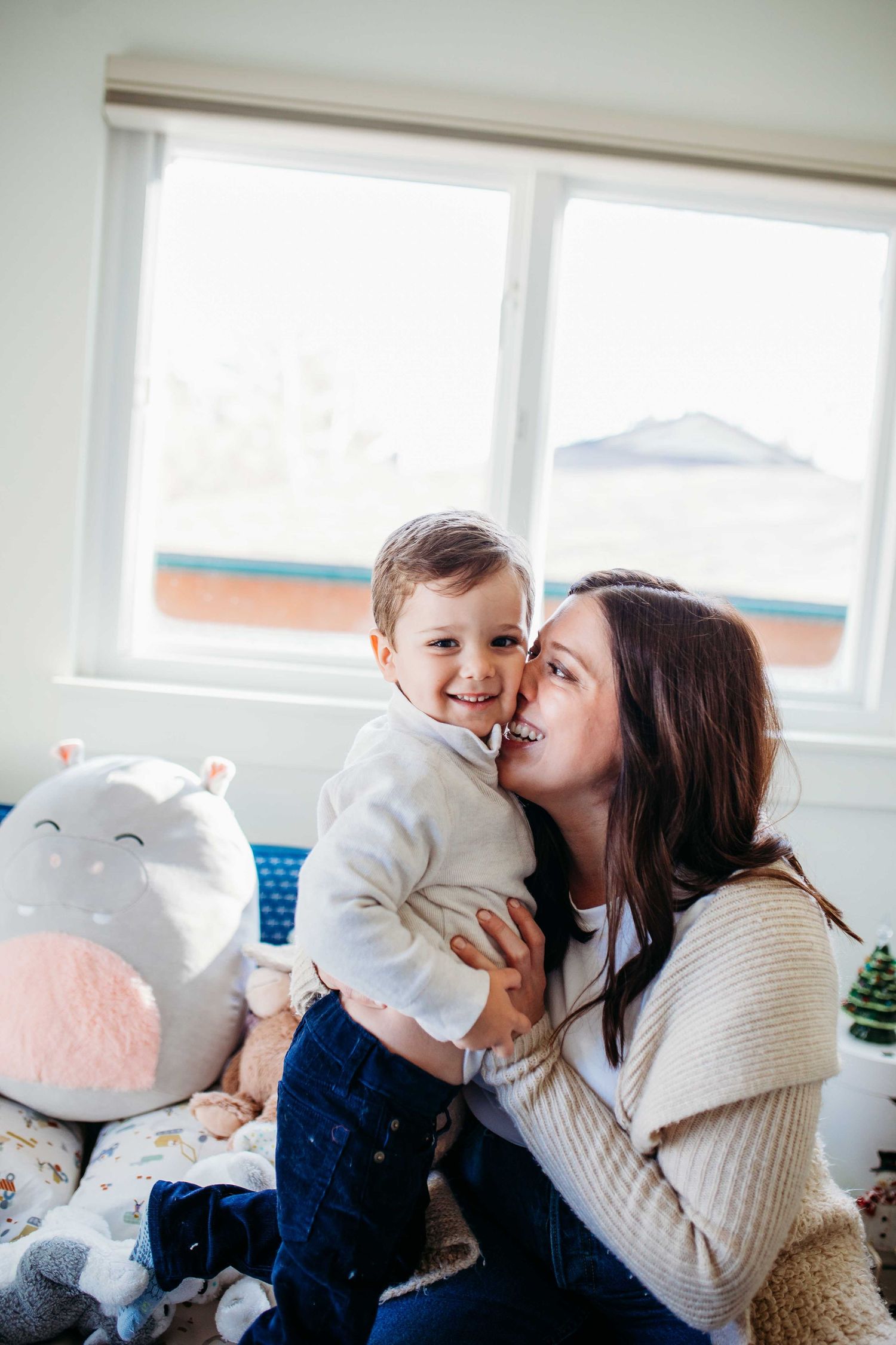 A parent and child share a tender moment while sitting together near a window with a stuffed animal.