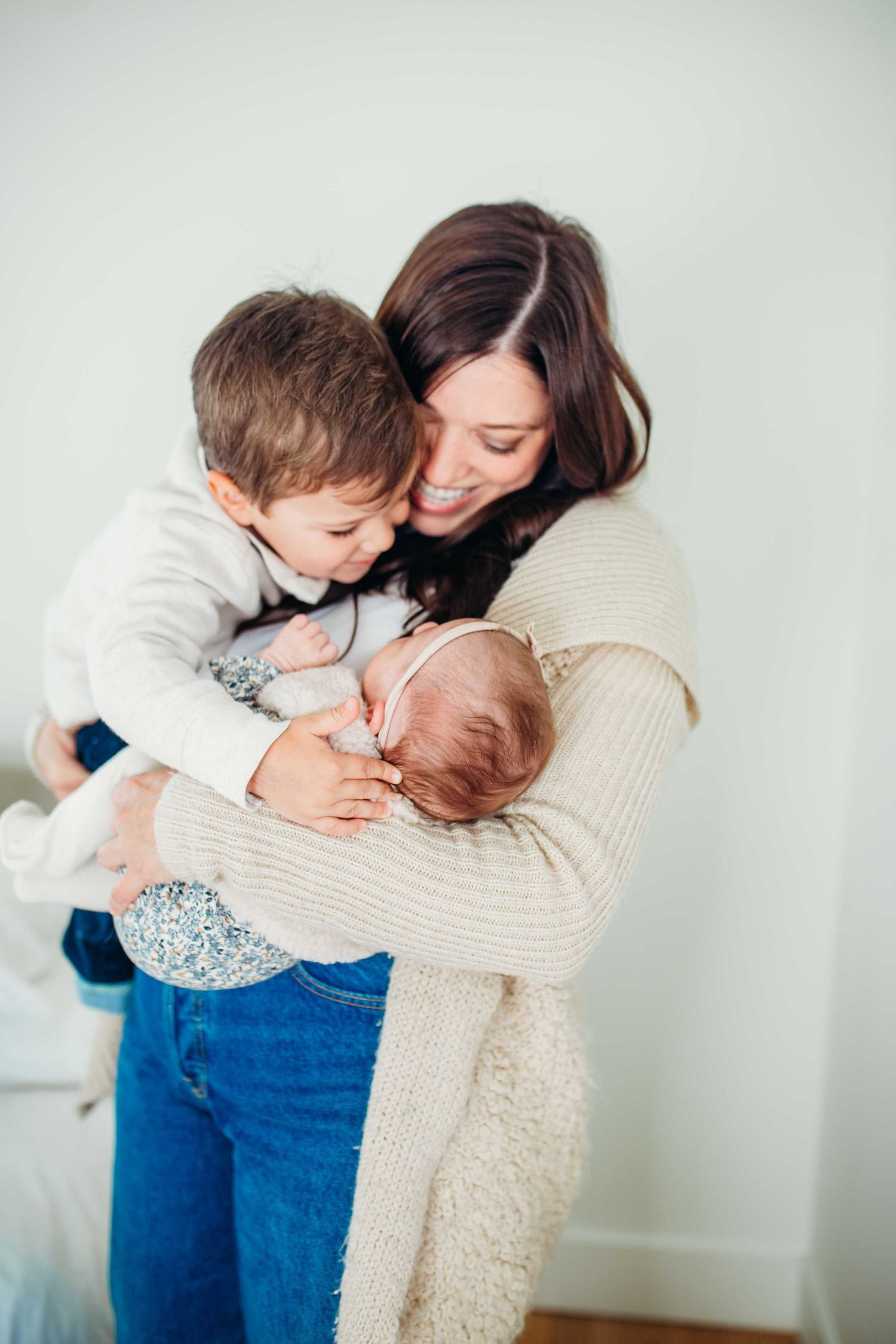 Caring parent in cream sweater embraces new baby and toddler with tenderness against white wall.