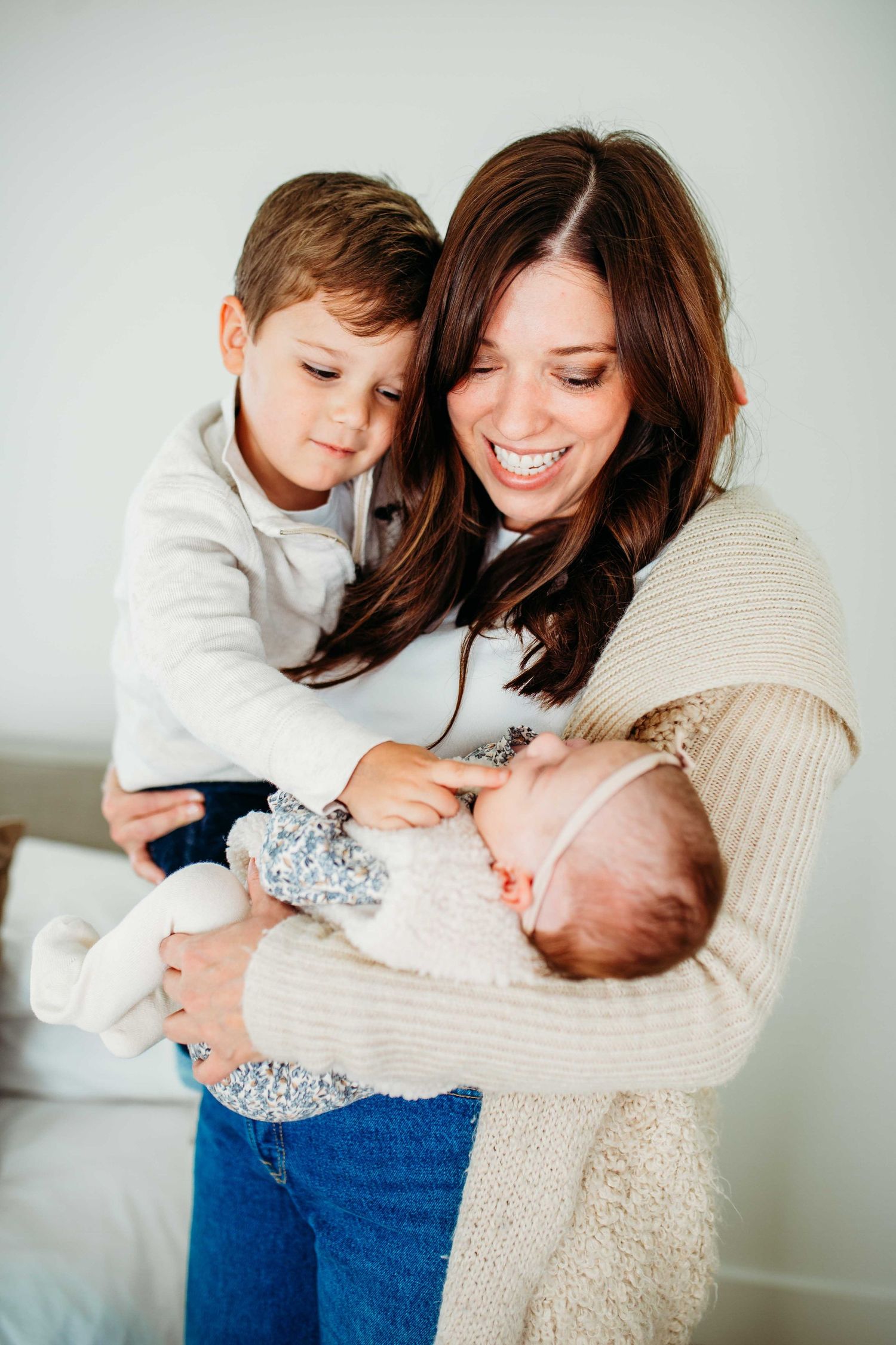 Woman in beige sweater and jeans holding newborn baby while young child leans in to look at sibling.