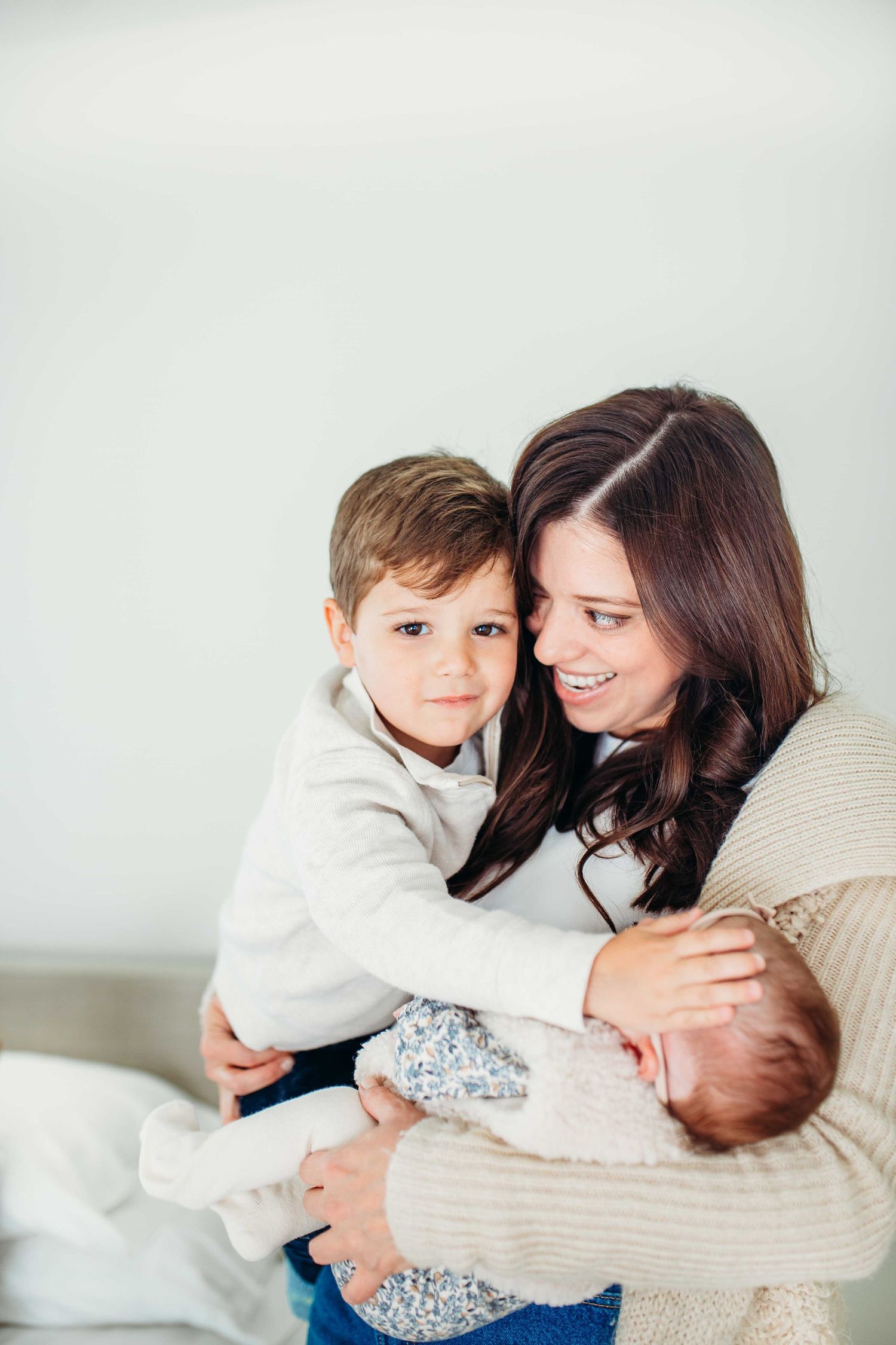 A loving family moment shows a mother in a beige sweater embracing two small children on a white background.