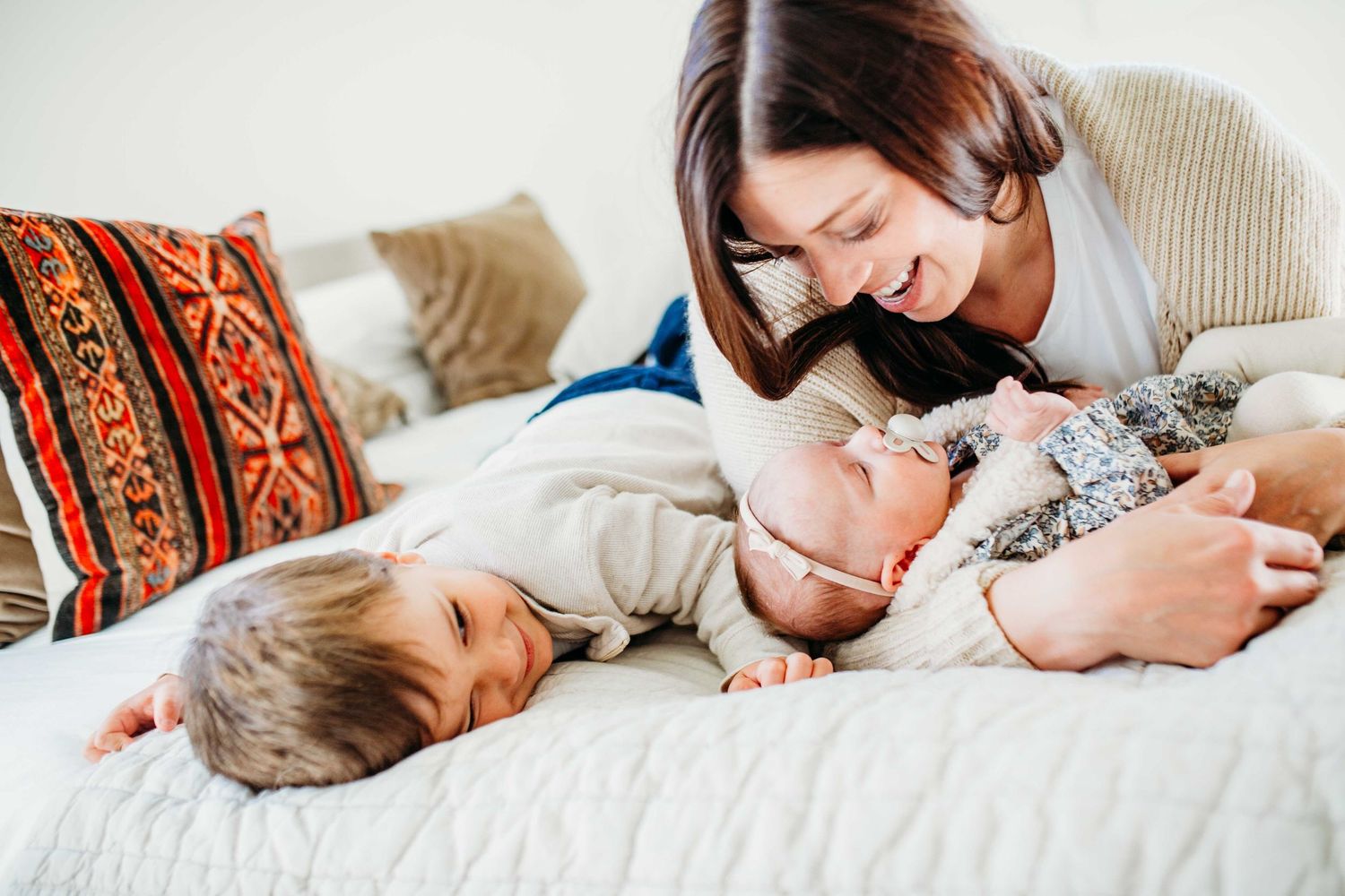 Two siblings lying side by side on a bed with decorative pillows while playing and laughing together.