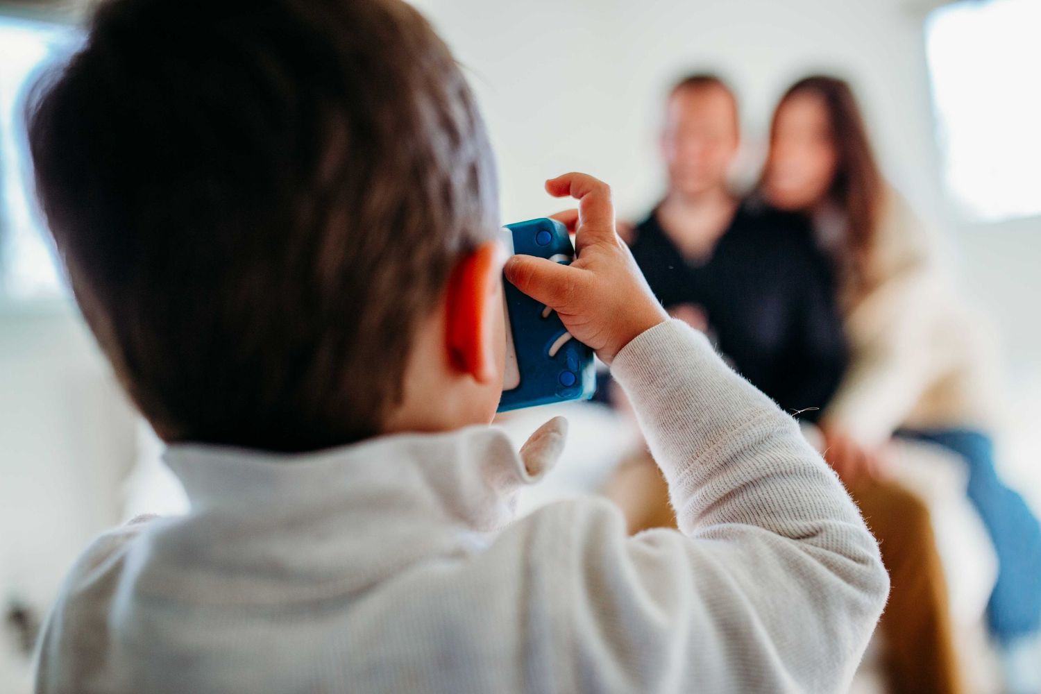 Child holding a blue toy camera and taking pictures indoors while facing towards blurred people in background.