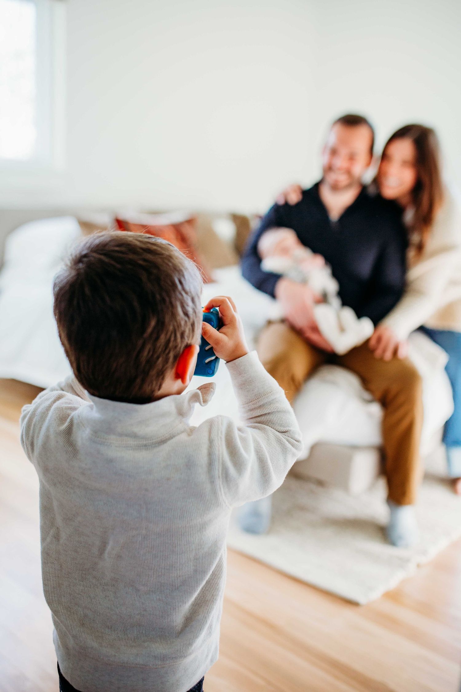Small child wearing a gray sweatshirt takes photos with a camera on hardwood floor while people sit in background.