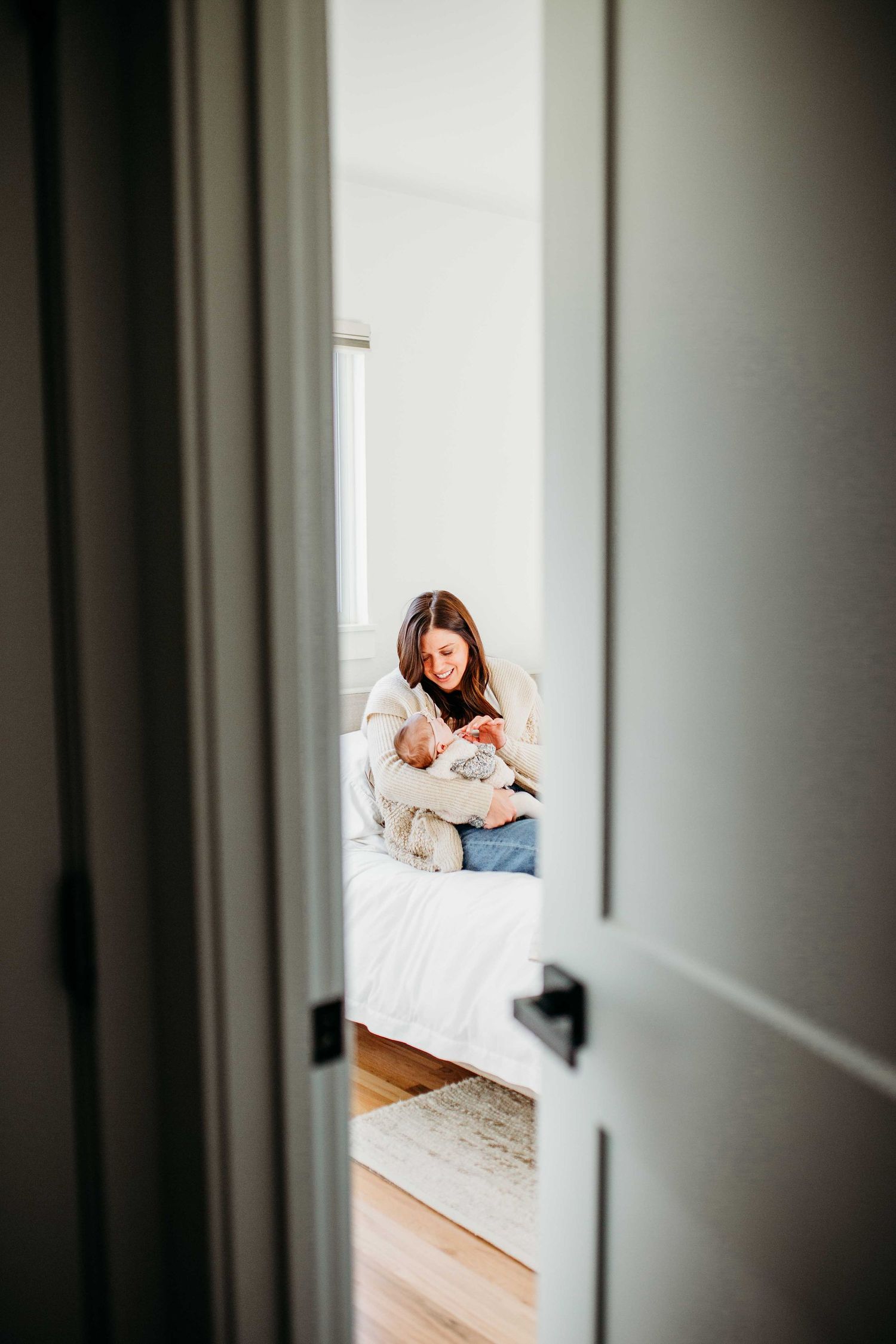 Through a doorway, a person in white sits on a bed holding a small baby in a cozy bedroom setting.