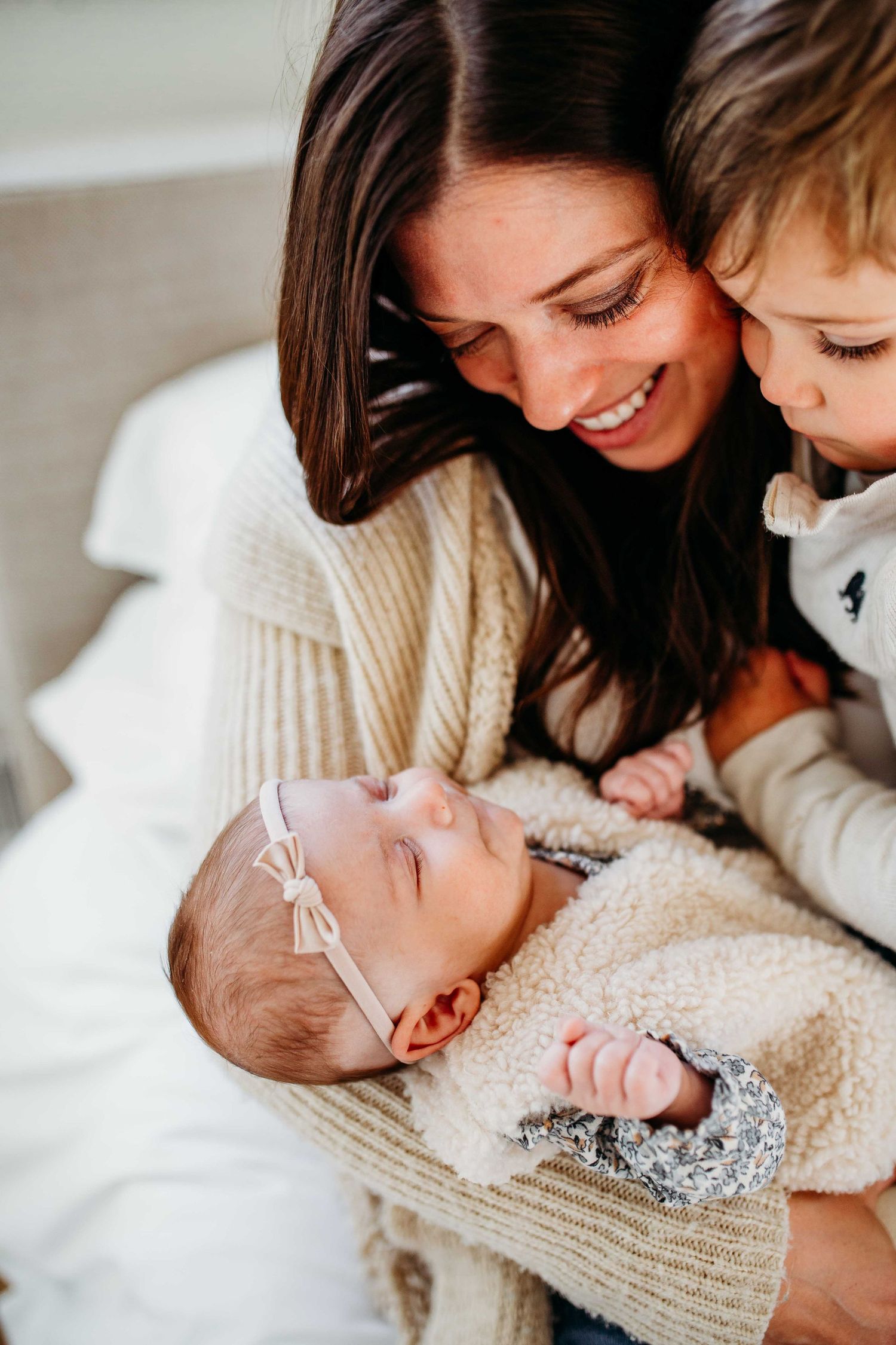 A sweet candid family moment with a newborn baby wearing a headband and siblings sharing tender smiles together.