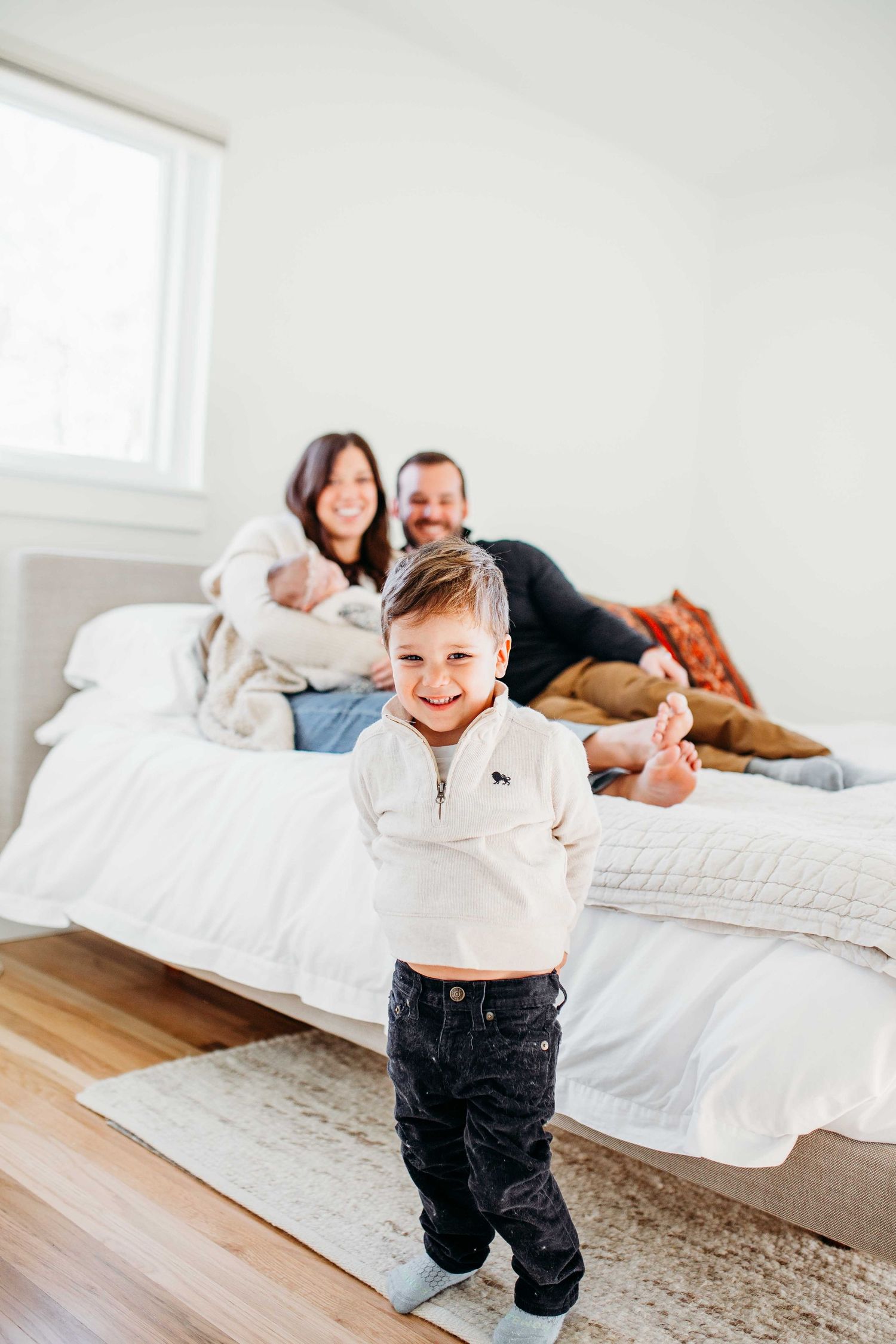 A joyful family moment as they relax together on a white bed in a bright, minimal bedroom with hardwood floors.