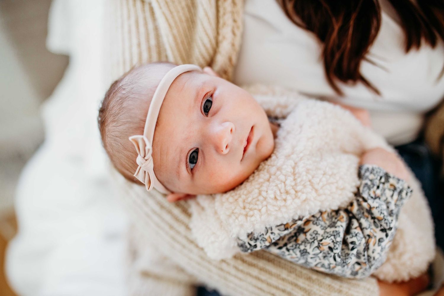 A baby wearing a white knit hat lies on soft bedding while looking at the camera.