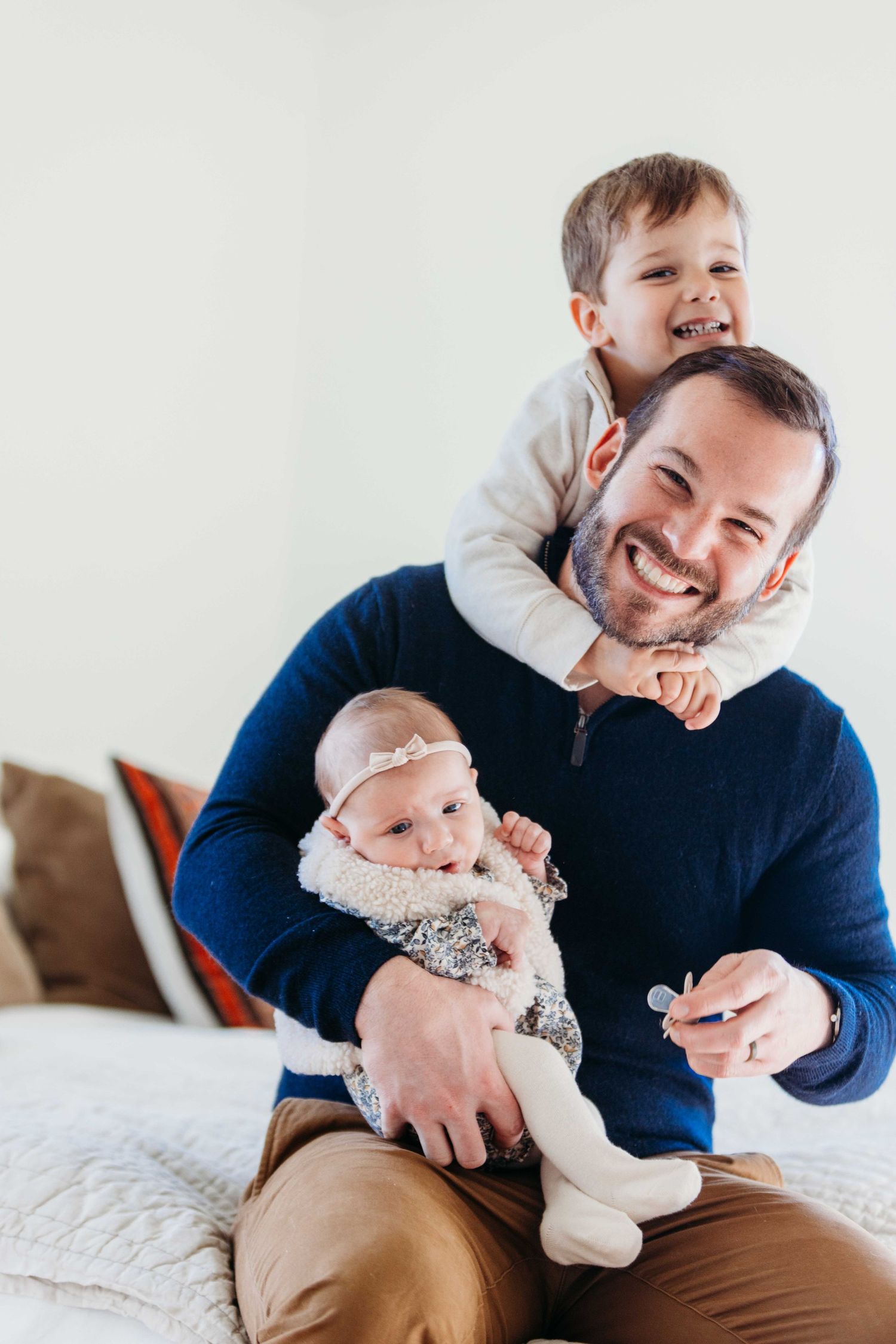 A beaming parent sits on a couch while holding an infant and playing with a toddler on their shoulders.
