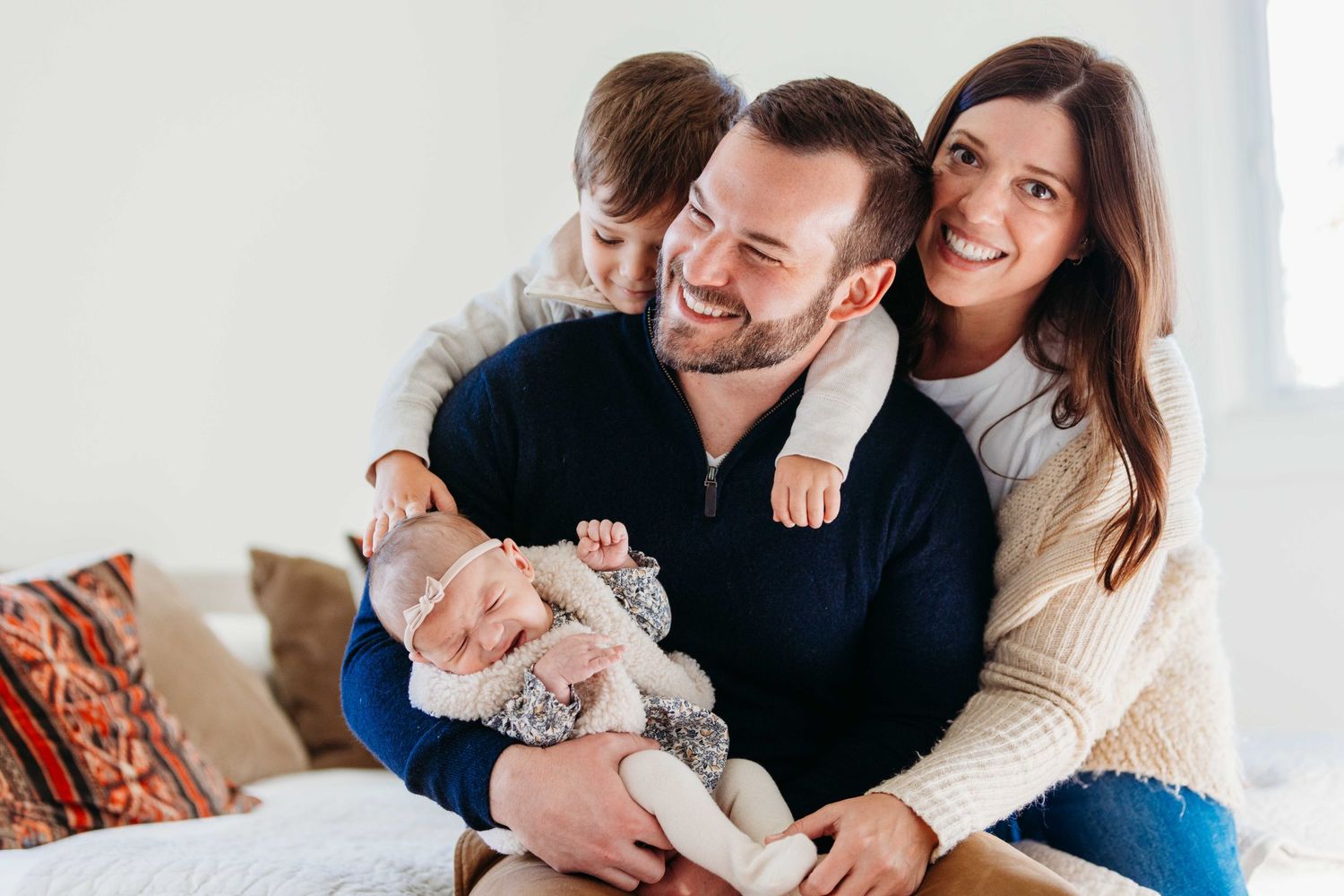 A smiling family relaxes together on a cozy bed with decorative pillows in a bright sunlit room.