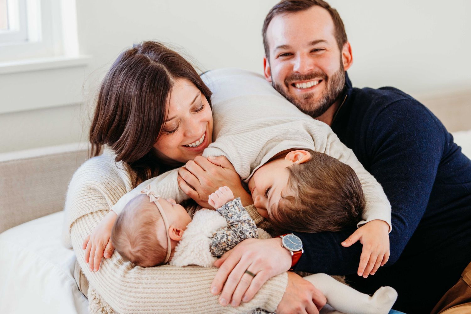 Family members share tender moments of affection and laughter while cuddling together on white bedding.