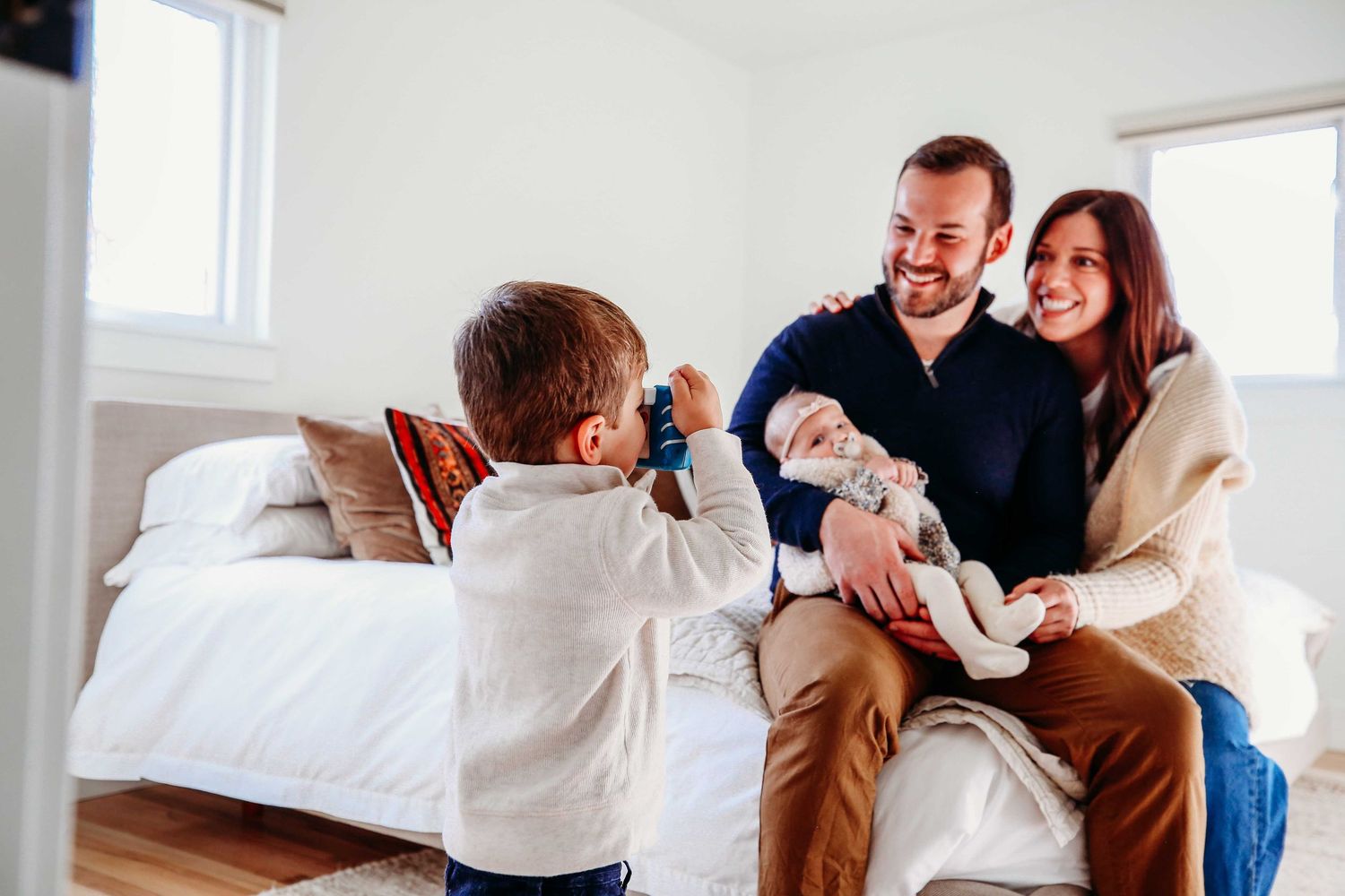 A young child takes a photo of a couple with an infant sitting together in a bright, minimalist room.
