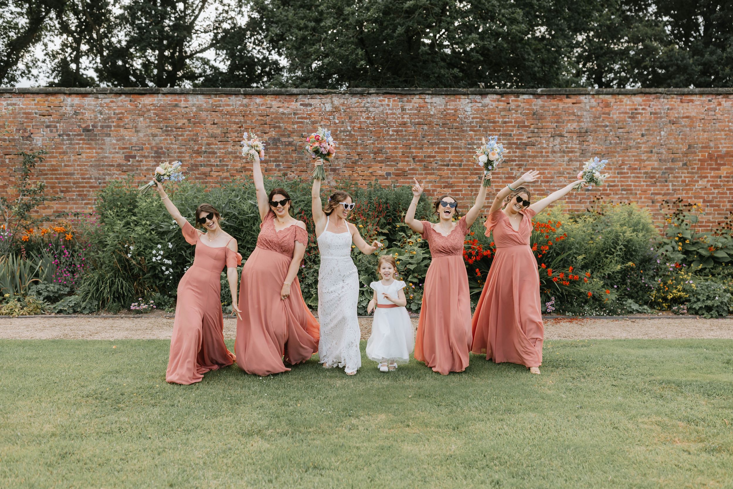 Wedding party in peach-colored bridesmaid dresses celebrate joyfully on lawn in front of brick garden wall.