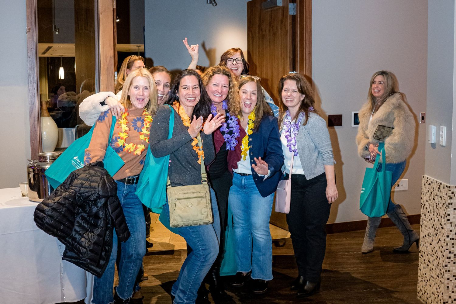 A group of enthusiastic women pose together at an indoor gathering, wearing casual attire and carrying bags.