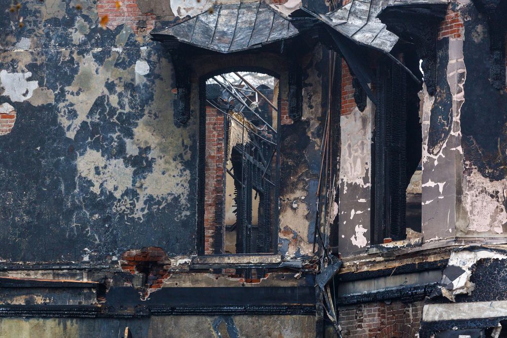 Weathered and charred window frame of a burned building with peeling paint and exposed brick walls.