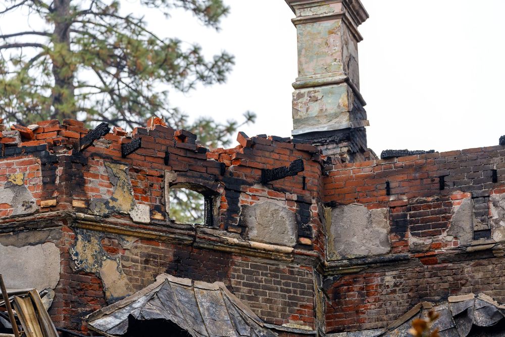 Crumbling brick wall and damaged chimney of an old ruined building against trees in background.