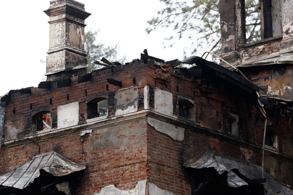 Damaged brick building with burned and deteriorating roof showing exposed structural elements and chimney.
