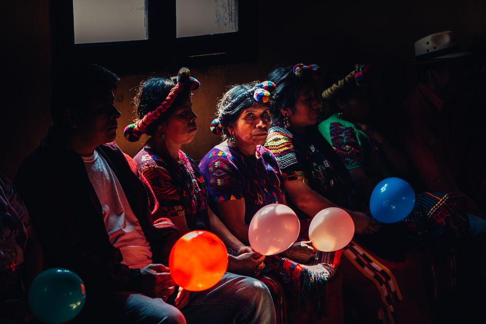 Several people sit together in a dark room holding illuminated colorful balloons that glow in red, orange and blue tones.