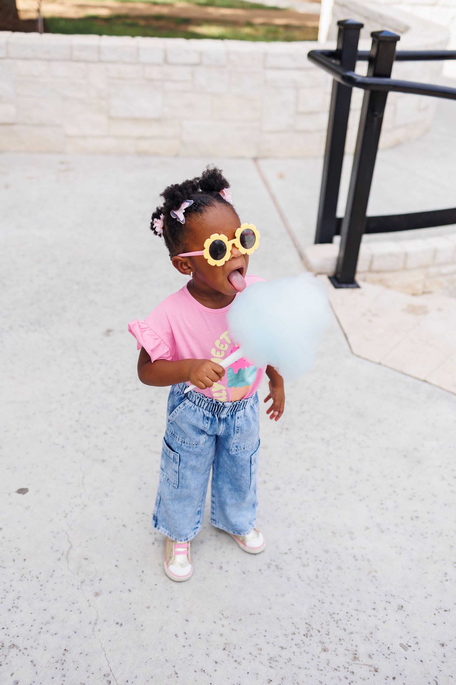 Small child wearing yellow sunglasses, pink shirt and denim jeans enjoys cotton candy at a first birthday party at Bella Cavalli